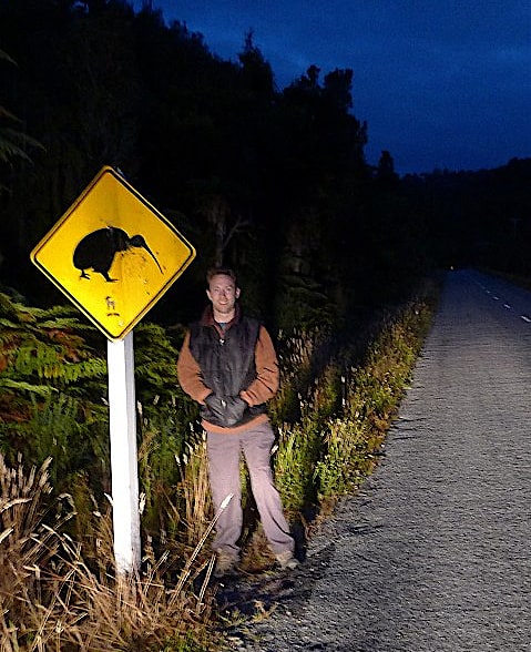 Chris Chalk in New Zealand standing next to a beware kiwi crossing the road sign, on the west coast of the south island near Okarito.